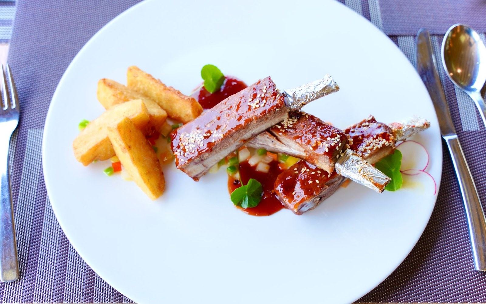 Glazed rib bones with sesame seeds, potato wedges, and diced vegetables on a white plate
